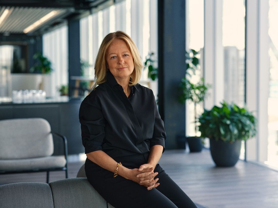 A senior professional in a black blouse seated in a bright modern office with plants and tall windows behind her.
