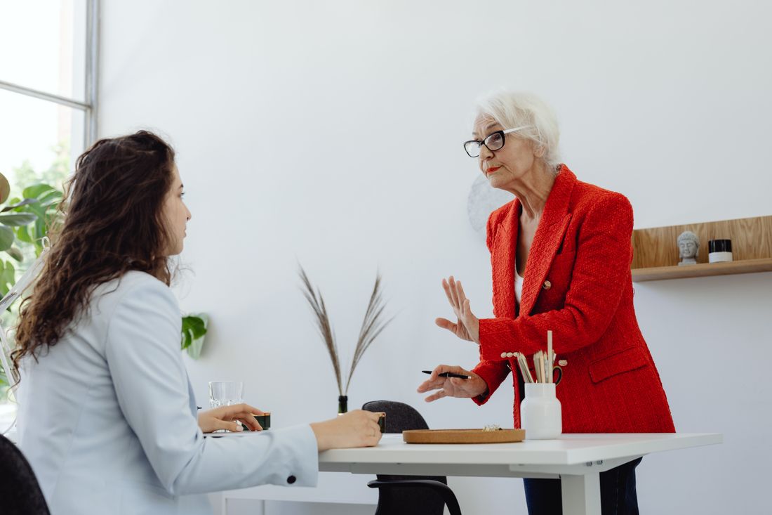Two professionals having a natural conversation at a plain office table while one listens and the other gestures.