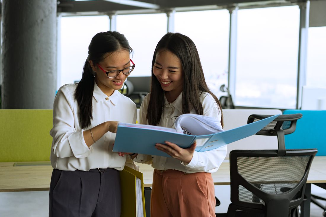 Two colleagues in business-casual attire reviewing a binder of documents together in a bright office.