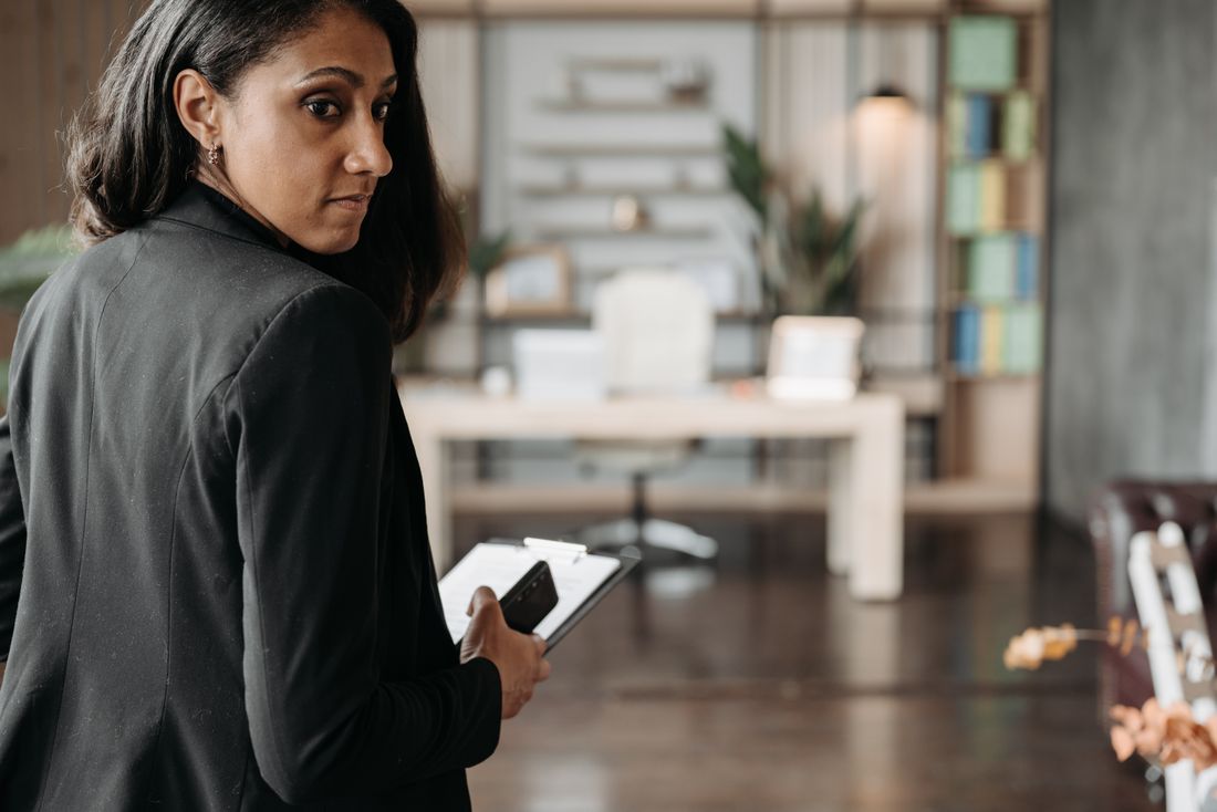 A businesswoman in a dark blazer glances back while standing in a modern office.