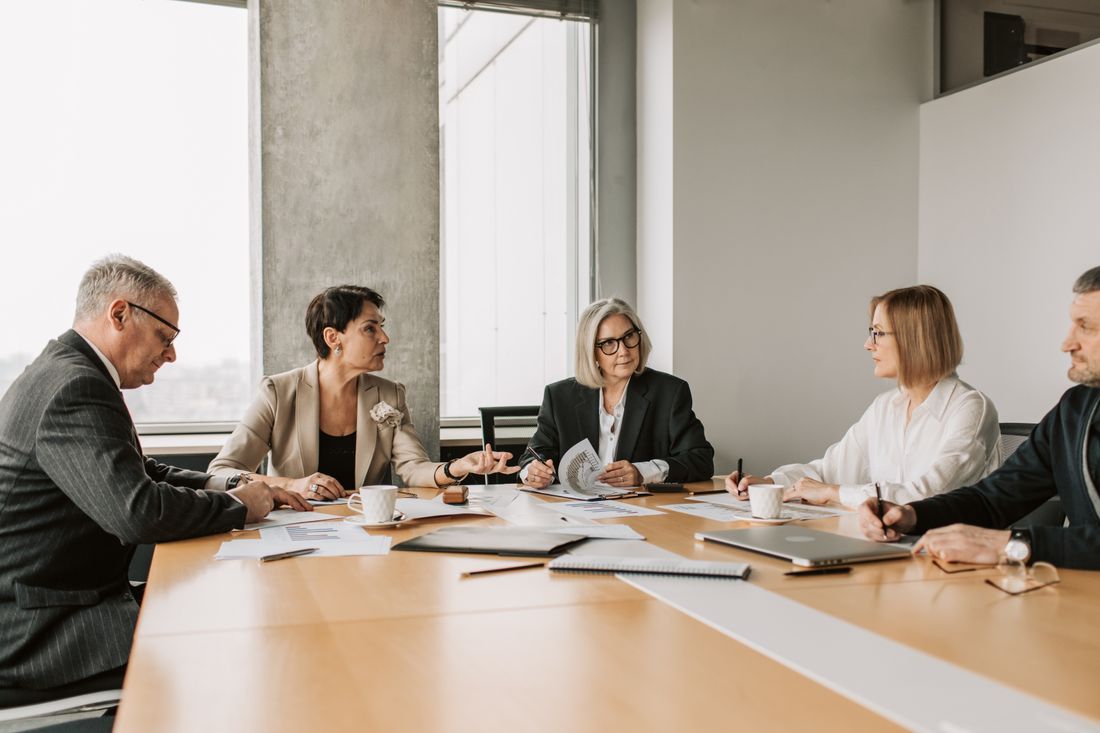Five senior professionals in business attire seated around a conference table in serious discussion, one woman mid-sentence and gesturing while others listen and take notes, papers and charts spread on the table.