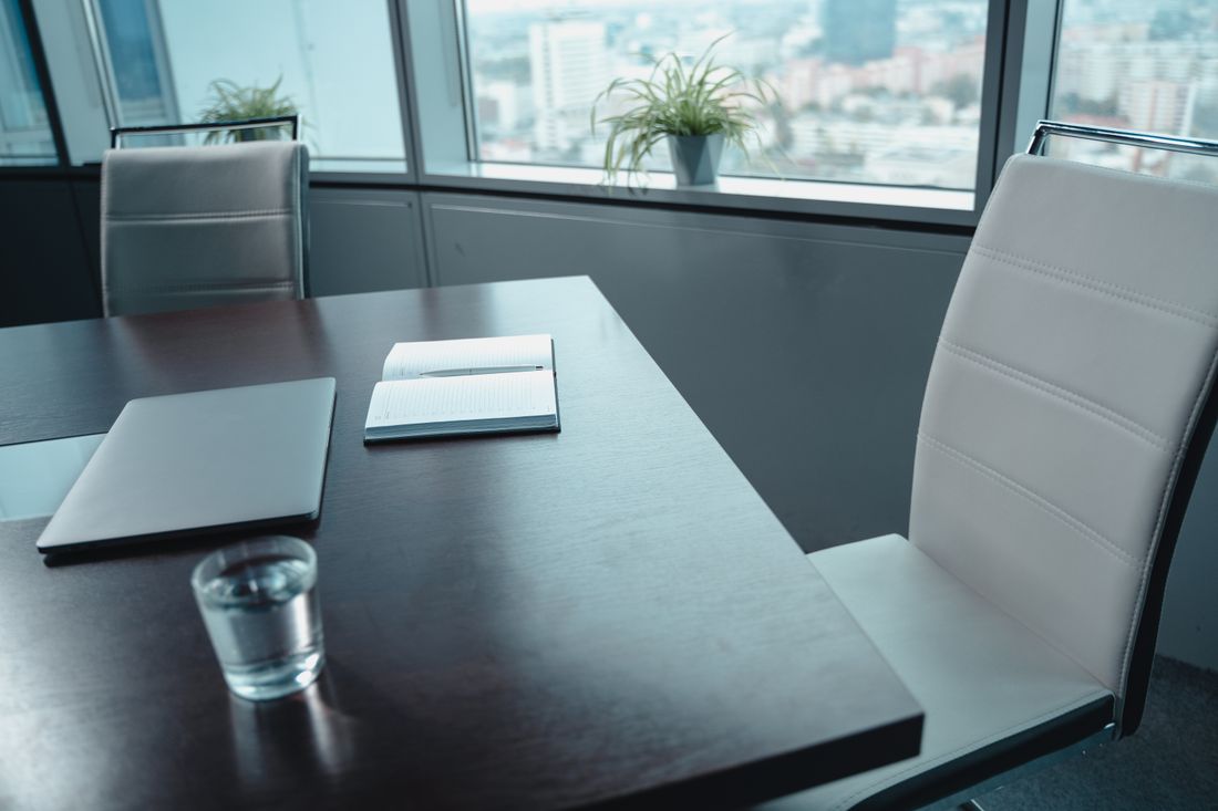 A quiet conference table corner with an open notebook and pen, a closed laptop, and a glass of water, shot at an angle with soft natural light and an empty chair in the foreground.