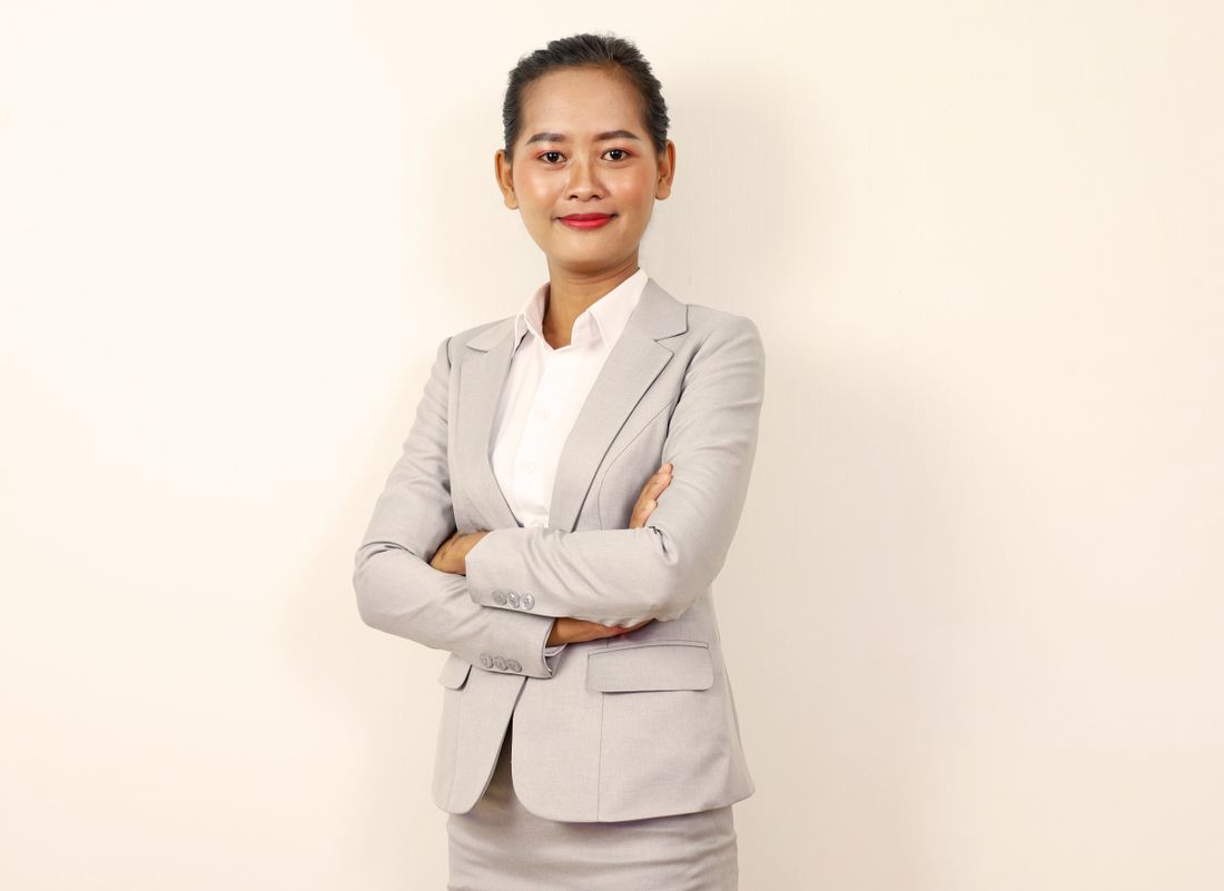 A woman in a gray business suit stands with arms crossed against a plain light backdrop, looking at the camera.