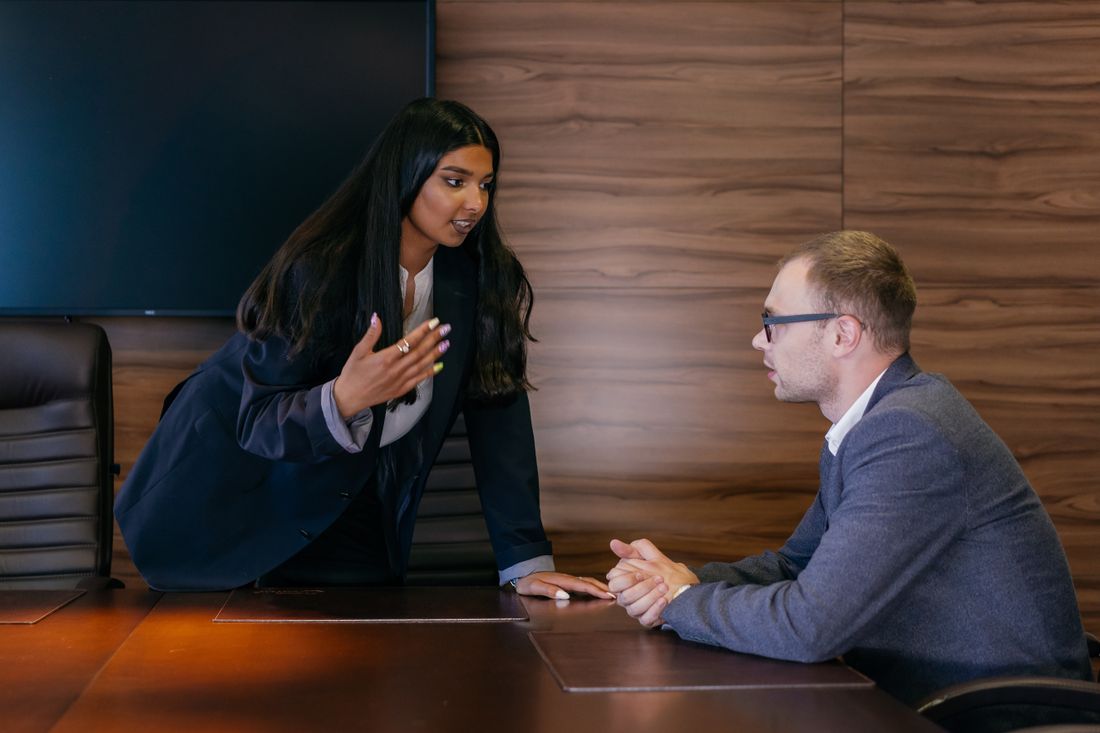 A woman leans across a conference table mid-gesture, talking to a seated colleague who listens with hands clasped.