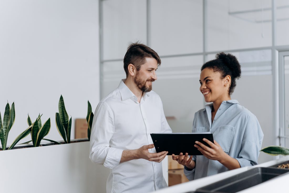 A man and woman stand near a plain wall with potted plants, smiling as they look at each other while he holds a tablet between them.