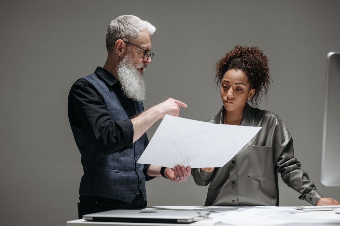 A bearded man points at a sketched diagram on a sheet of paper while a woman beside him looks on, standing at a table.