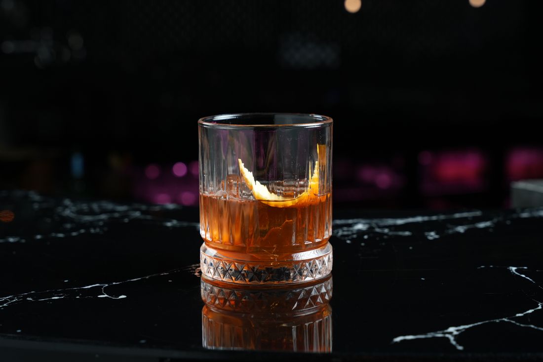 A single amber cocktail with an orange peel garnish in a cut-crystal rocks glass, backlit against a dark bar backdrop on a black marble counter with visible condensation.
