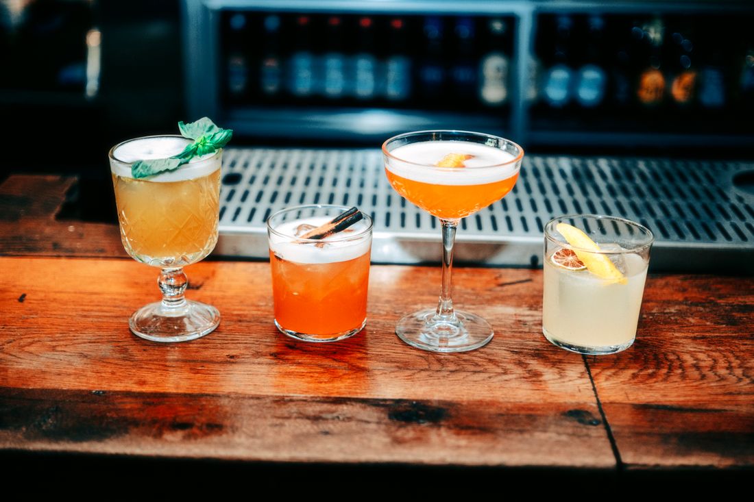 Four different cocktails lined up on a wooden bar counter, each in its own glass with a citrus or herb garnish, set against a dark backlit bottle display.