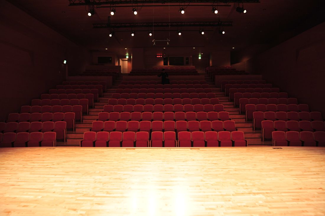 An empty theater auditorium with rows of vacant seats rising up from a bare wooden stage, lit warmly before an event.