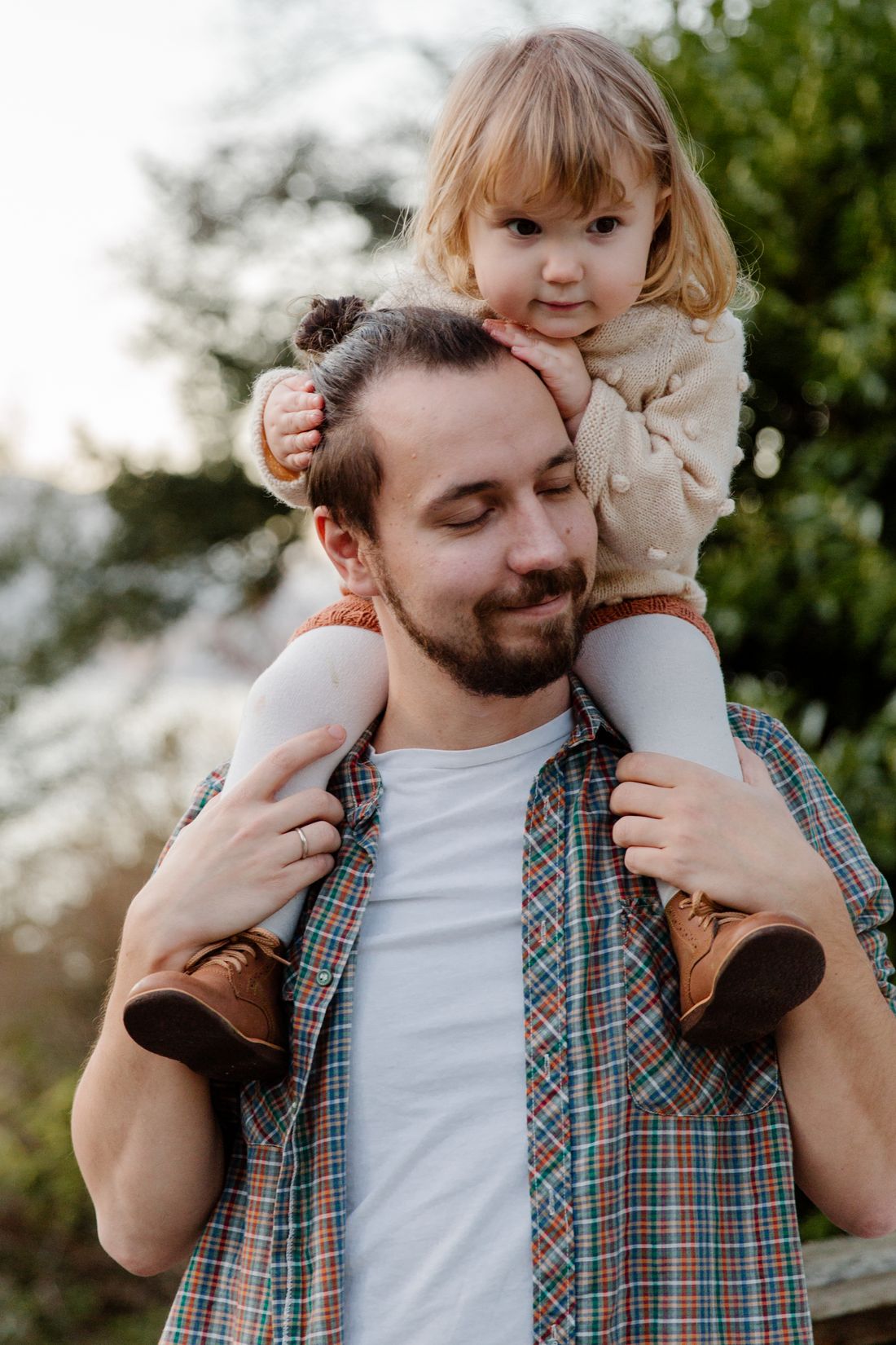A father carries his young daughter on his shoulders outdoors, both smiling, with green trees in the background.