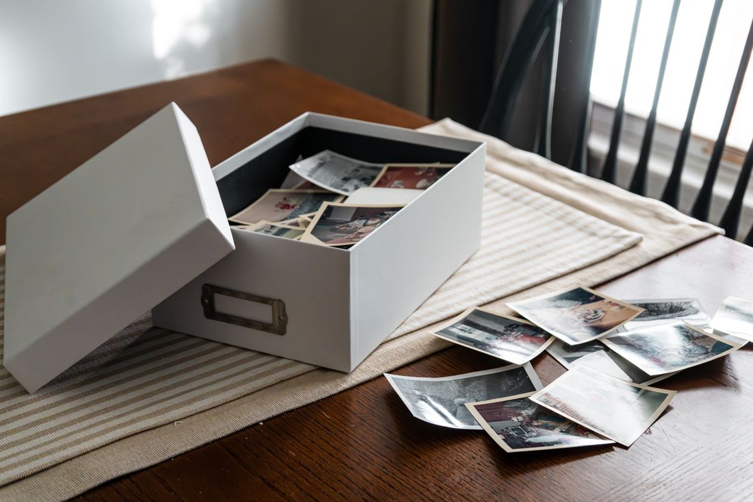 An open box of old printed photographs spilling across a wooden table, the kind of physical archive a company anniversary piece has to work with.