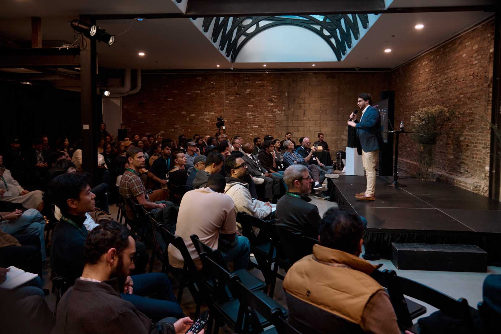 A wide shot of a speaker on stage addressing a full seated audience in a brick-walled venue.