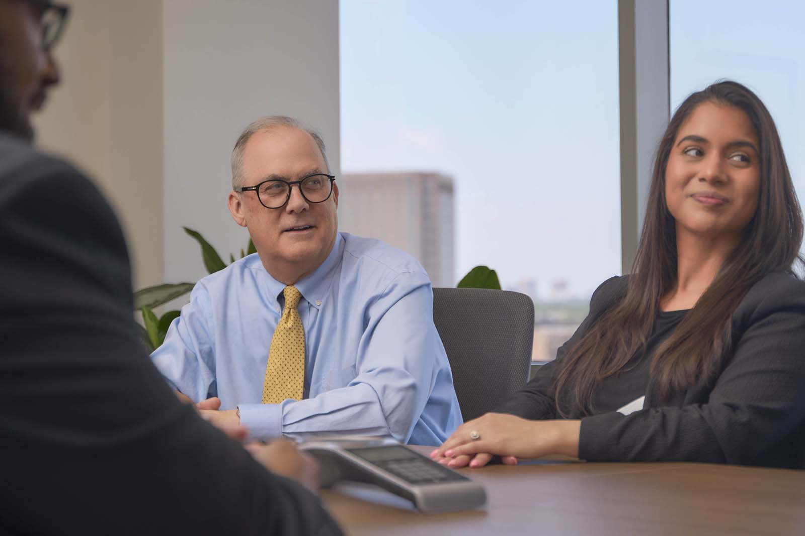 Two men and a woman conferring at a conference table with a speakerphone and the NYC skyline behind them