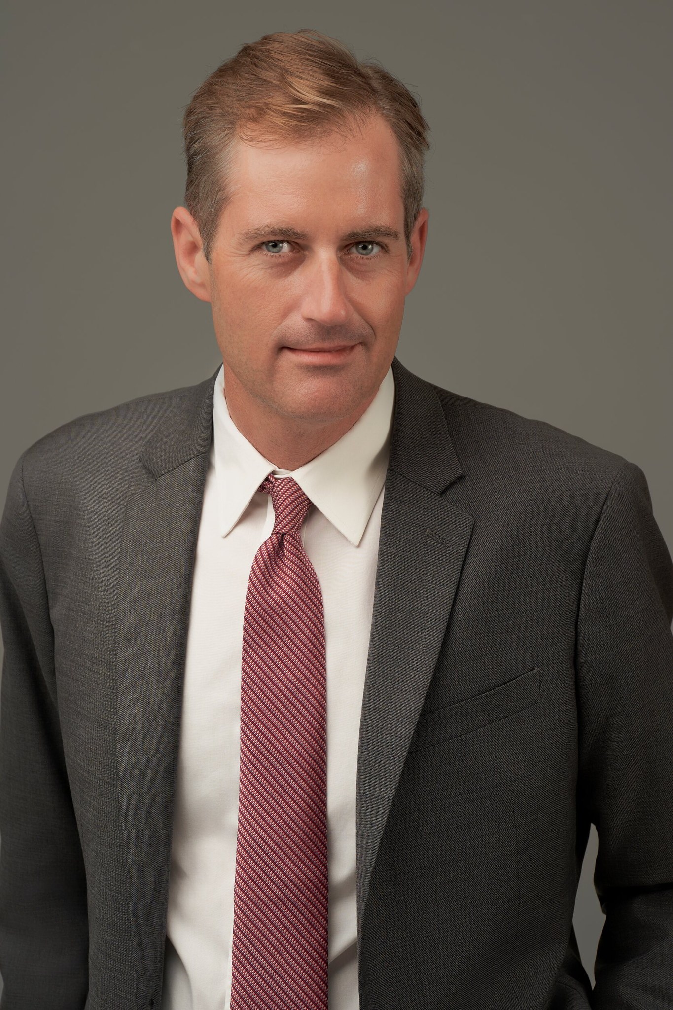 Man with short blonde hair in dark grey suit and maroon striped tie, professional studio headshot.