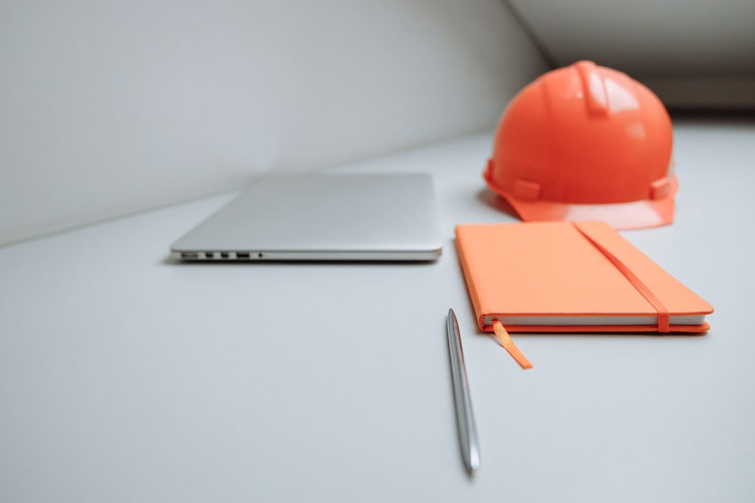 An orange hard hat resting on a desk next to a laptop, notebook, and pen.