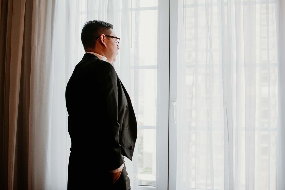 A person in a dark suit seen from the side and behind, standing at a large window looking out toward a blurred city view, natural daylight.
