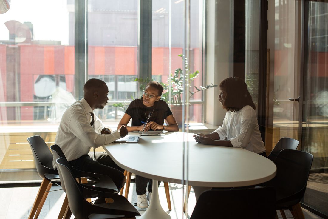 Three colleagues seated at a table mid-discussion inside a glass-walled meeting room, seen from outside the glass with daylight from office windows.
