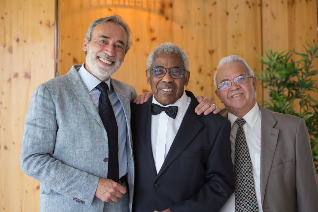 Three men in formal attire, two in suits and one in a tuxedo with a bow tie, stand close together with arms around each other, smiling warmly.