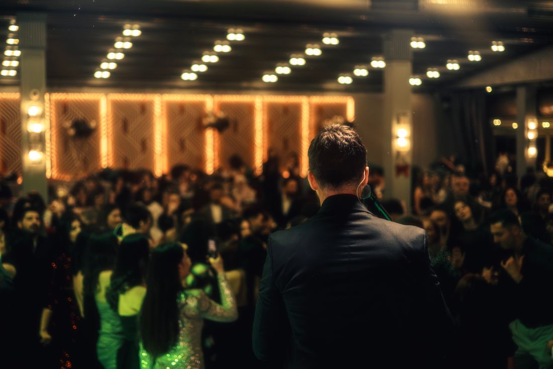 A man in a dark suit stands with his back to the camera facing a crowded, warmly lit room of guests in evening wear at a formal evening event.