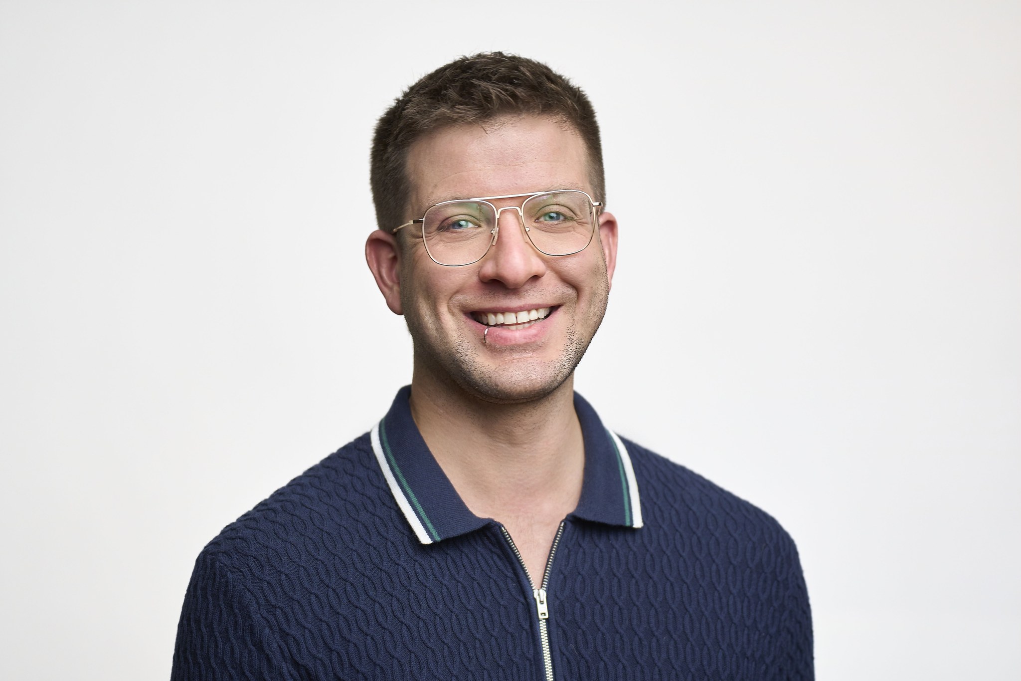 Smiling man with glasses in navy cable-knit polo poses for headshot against white background.