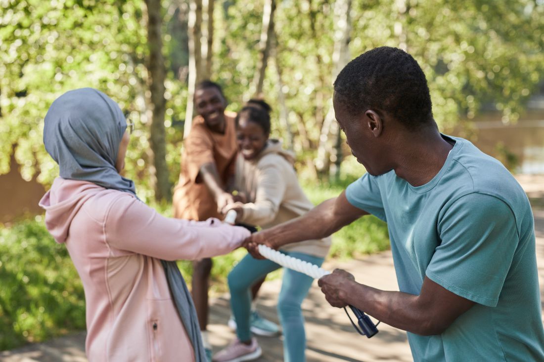 A group of coworkers in casual clothes laughing mid-tug-of-war on a sunny grass field, gripping a rope together outdoors.