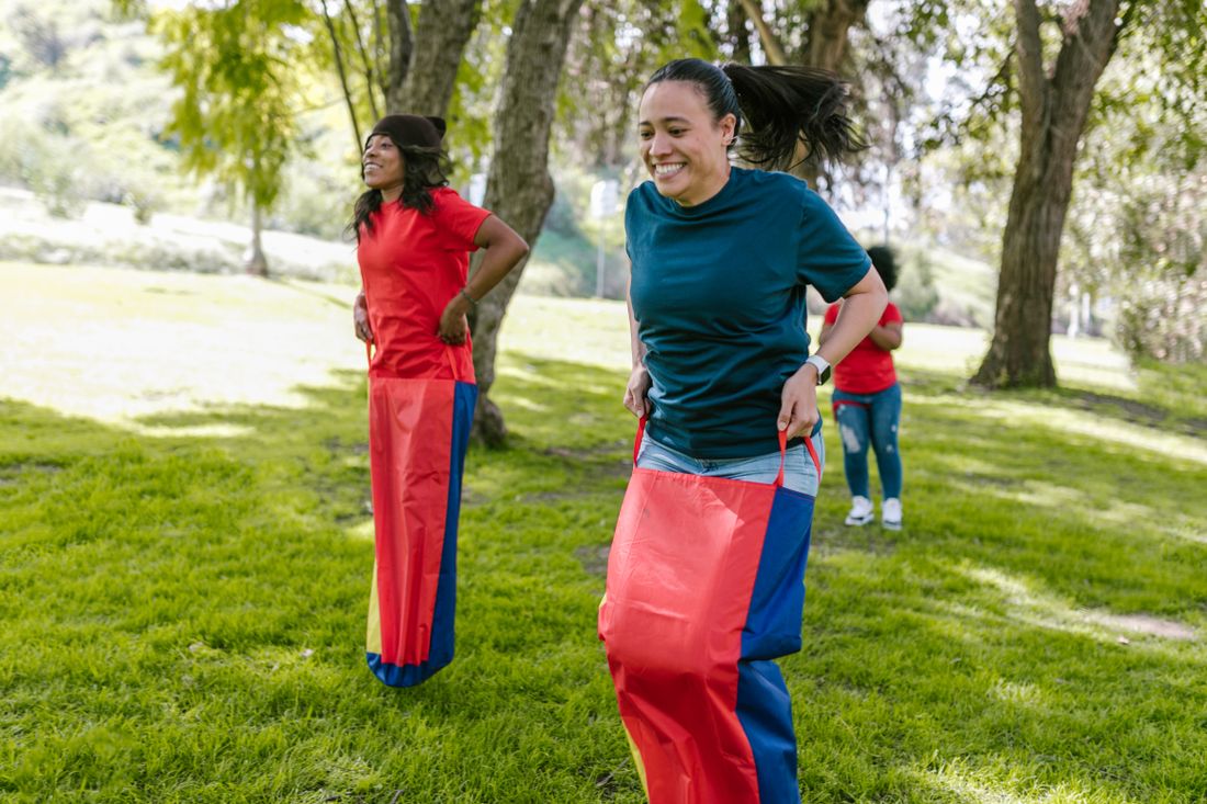 A small group of coworkers in casual clothes mid-motion during an outdoor sack race in a park, laughing and hopping across the grass.