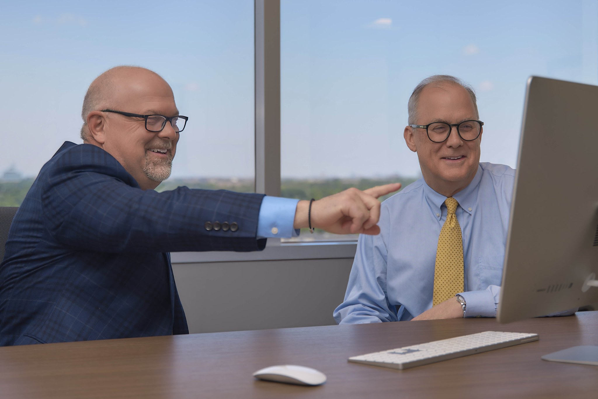 Two men at an office desk, one smiling and pointing toward a computer monitor.