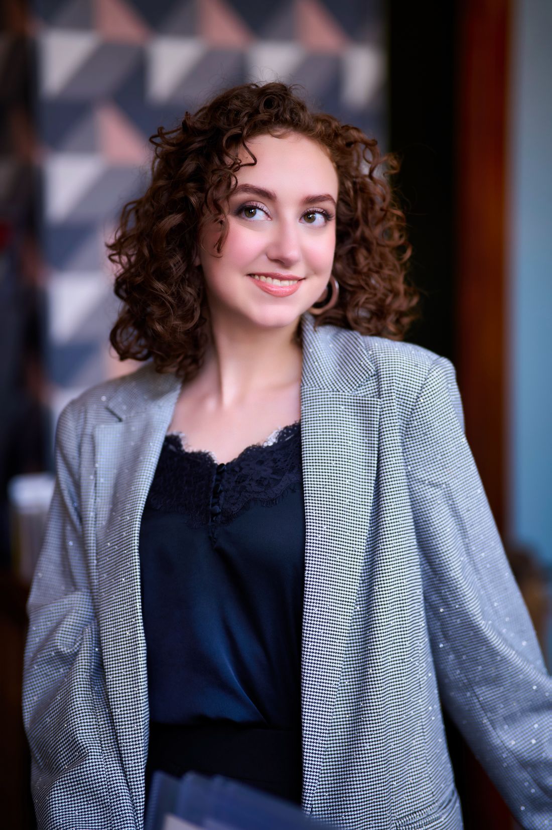 A woman in a smart-casual blazer smiles warmly in a softly lit indoor setting.