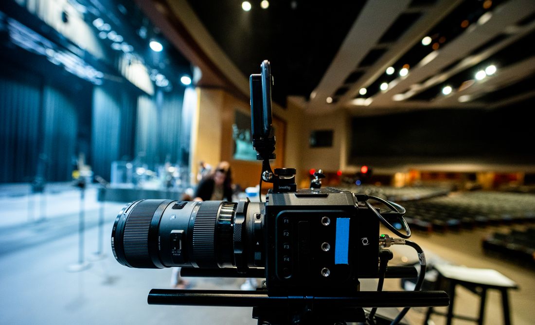 A single professional video camera locked off on a tripod near an empty stage, framed and ready before a program starts.