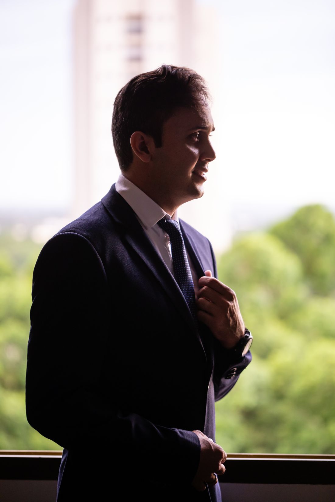 A business-professional man in a suit and tie standing indoors by a window, adjusting his tie in natural daylight with a soft green view behind him.