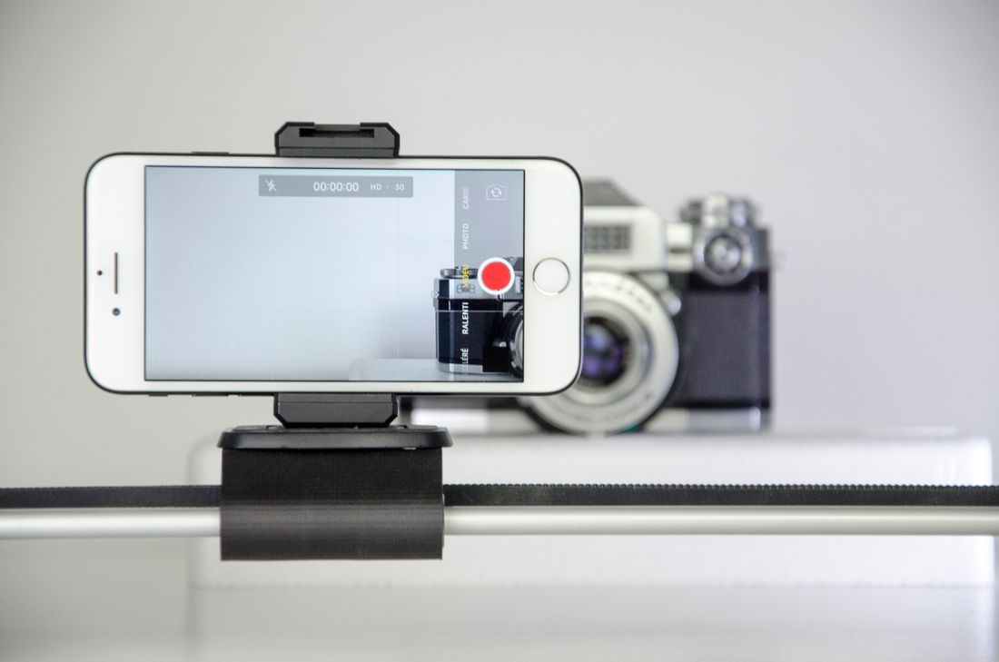 A smartphone mounted on a small rig records footage with a vintage camera visible out of focus behind it.