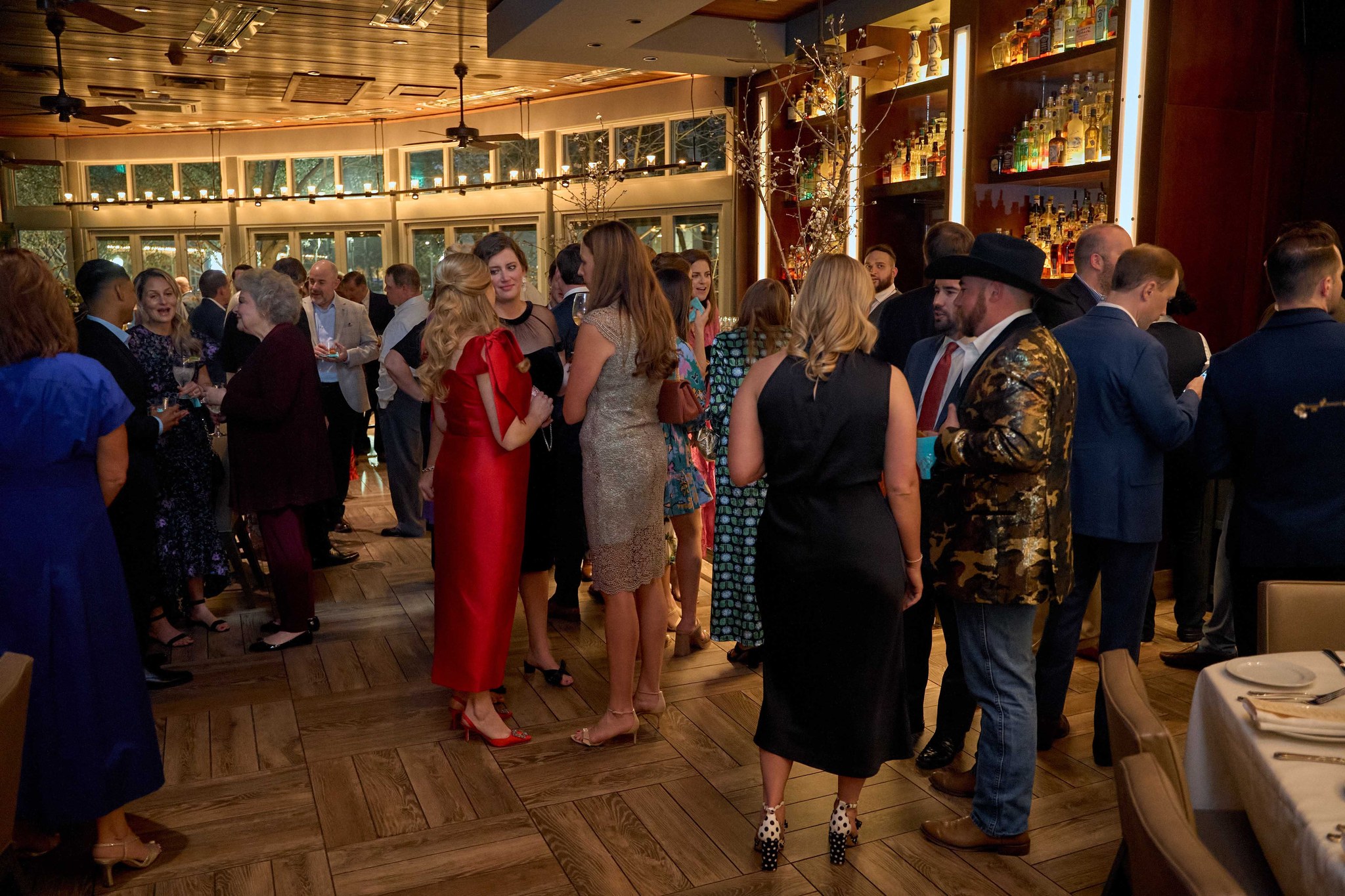Crowded restaurant dining room with guests in formal attire mingling near well-stocked bar.
