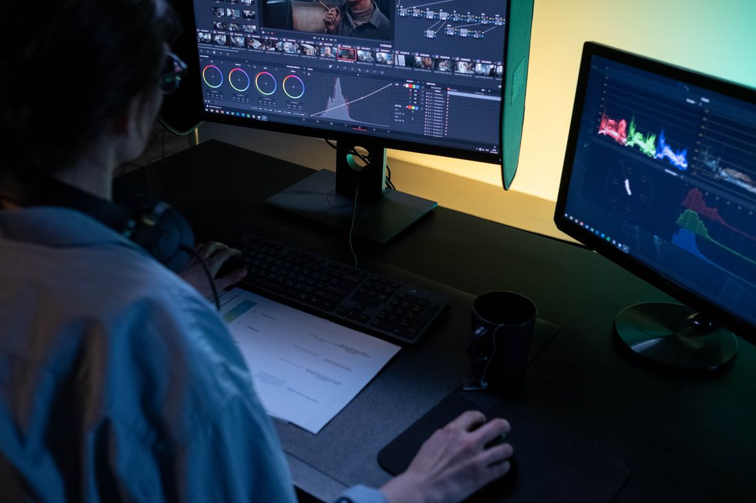 A video editor viewed from behind at a dual-monitor workstation, headphones around their neck, working a dense color-graded timeline packed with short clips on one screen and a scopes panel on the other.
