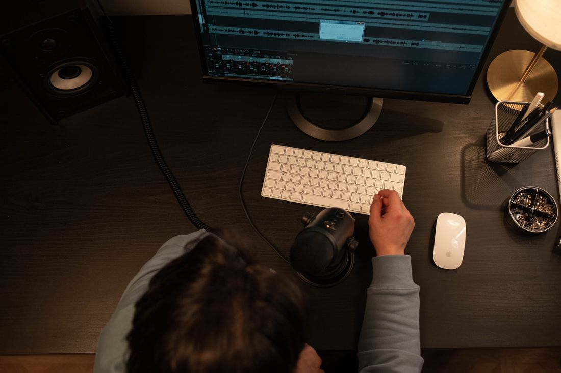 Overhead view of an editor's hand near a keyboard beside a monitor showing a dense, multi-clip video editing timeline, desk lamp casting warm light across the workspace.