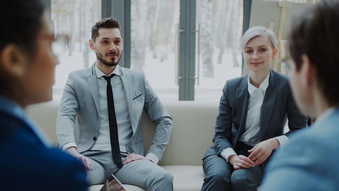 A man in a light gray suit and a woman in a dark gray blazer sit composed and poised together, facing the camera in a bright office.