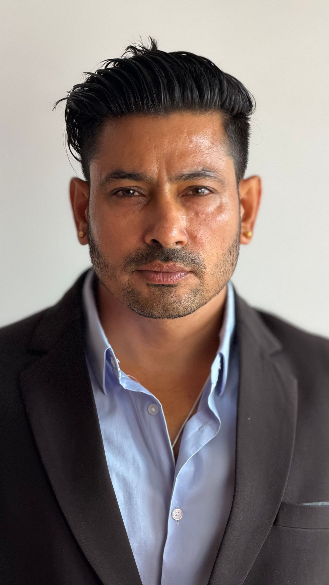 Head-and-shoulders portrait of a man in a dark suit jacket against a plain neutral wall, looking directly at the camera.
