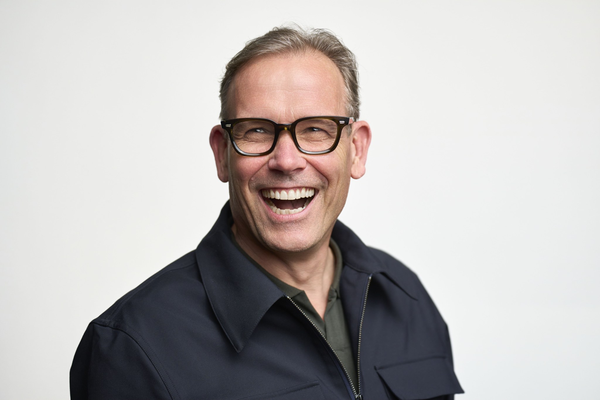 Older man with glasses laughing heartily in dark jacket over green shirt, approachable headshot.