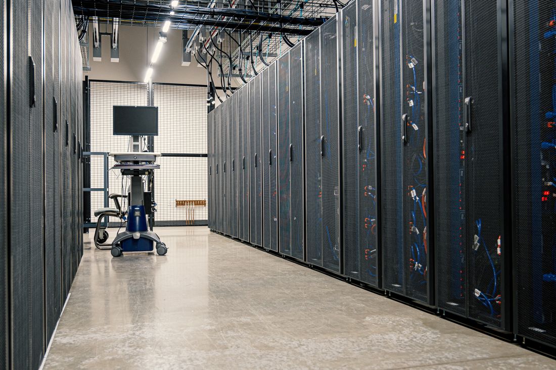A wide aisle between tall server racks with cabling visible, receding into the distance in a technical facility, no people in frame.
