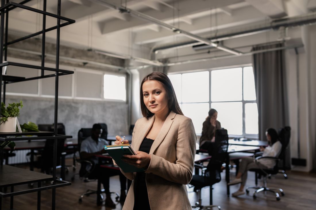 A woman in a blazer stands holding a notebook and pen in a busy open-plan office, with colleagues working at desks blurred in the background.