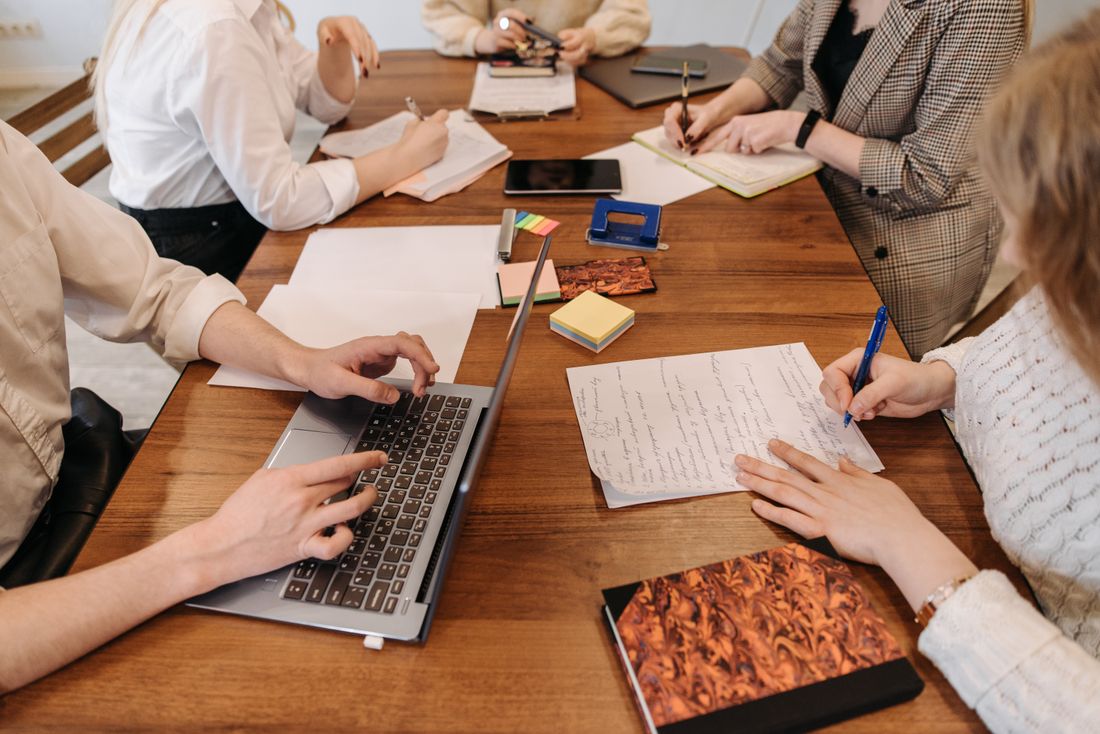 Several colleagues seated closely around a wooden table, hands filling a page with notes, typing on a laptop, and marking up papers scattered with sticky notes.