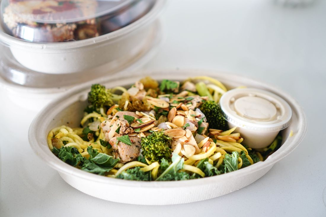 A plain fiber takeout bowl filled with prepared pasta, broccoli, and almonds, shot close and straight on with a small unlabeled sauce cup, neutral counter background