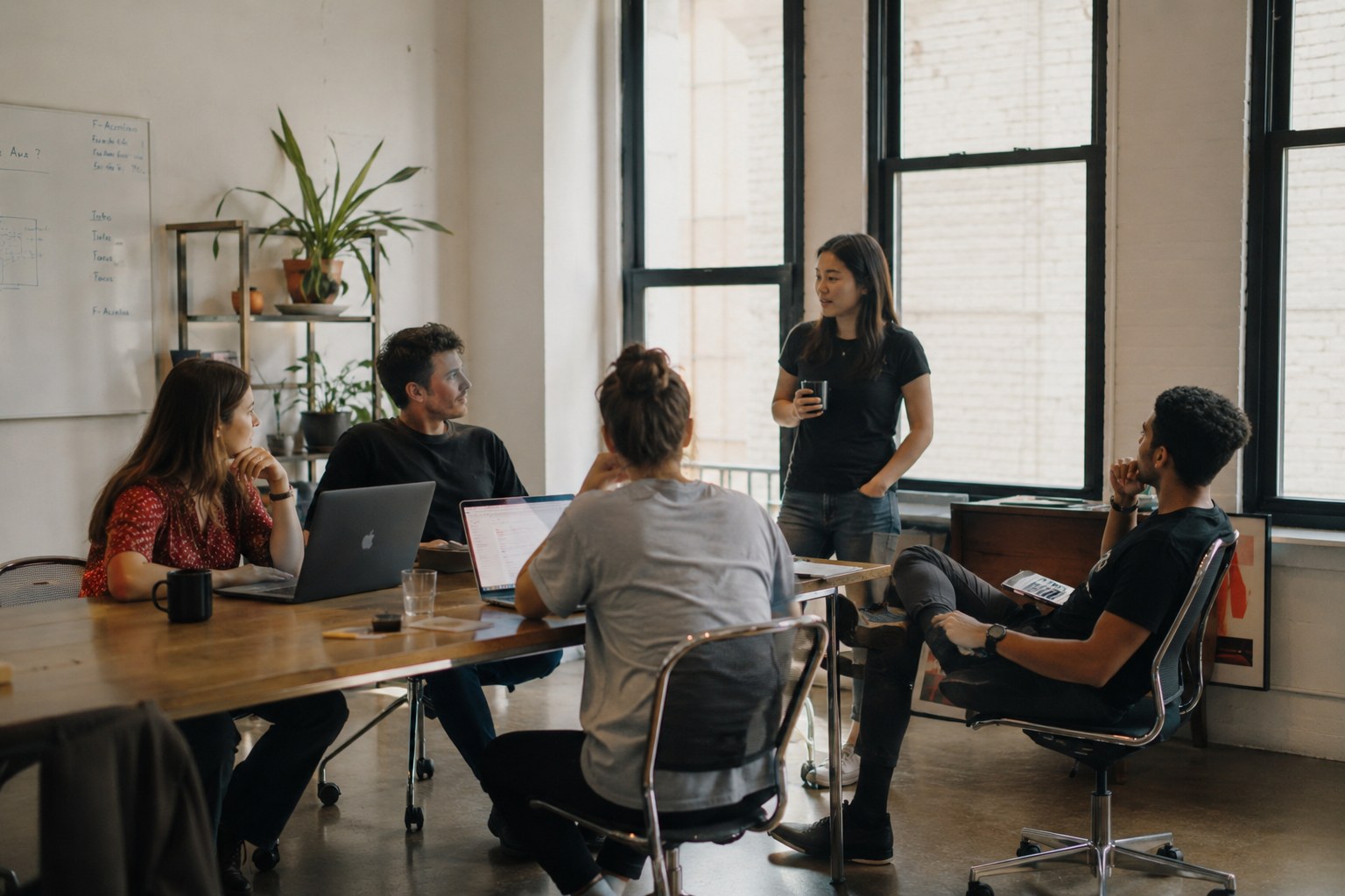 A group of developers gather around a table with laptops and whiteboard for discussion.