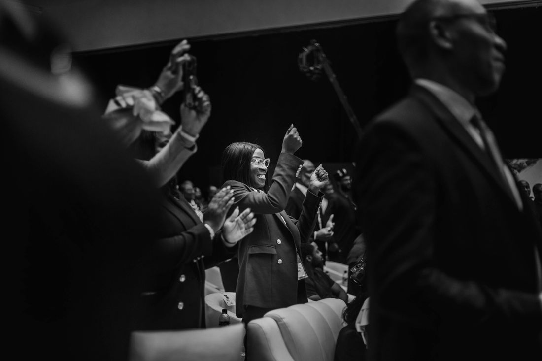 A group of well-dressed attendees applauding at a formal indoor event, in elegant black and white.