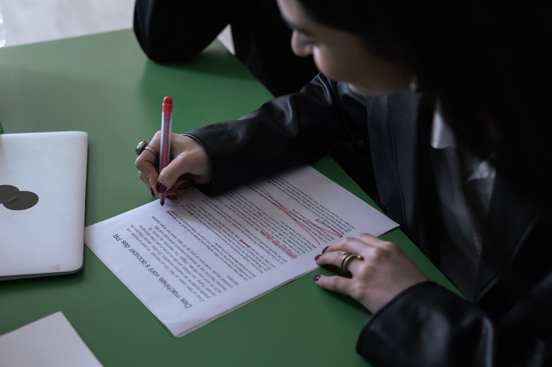 A hand marks up a printed document with a red pen next to an open laptop, reviewing edits before sending feedback back.