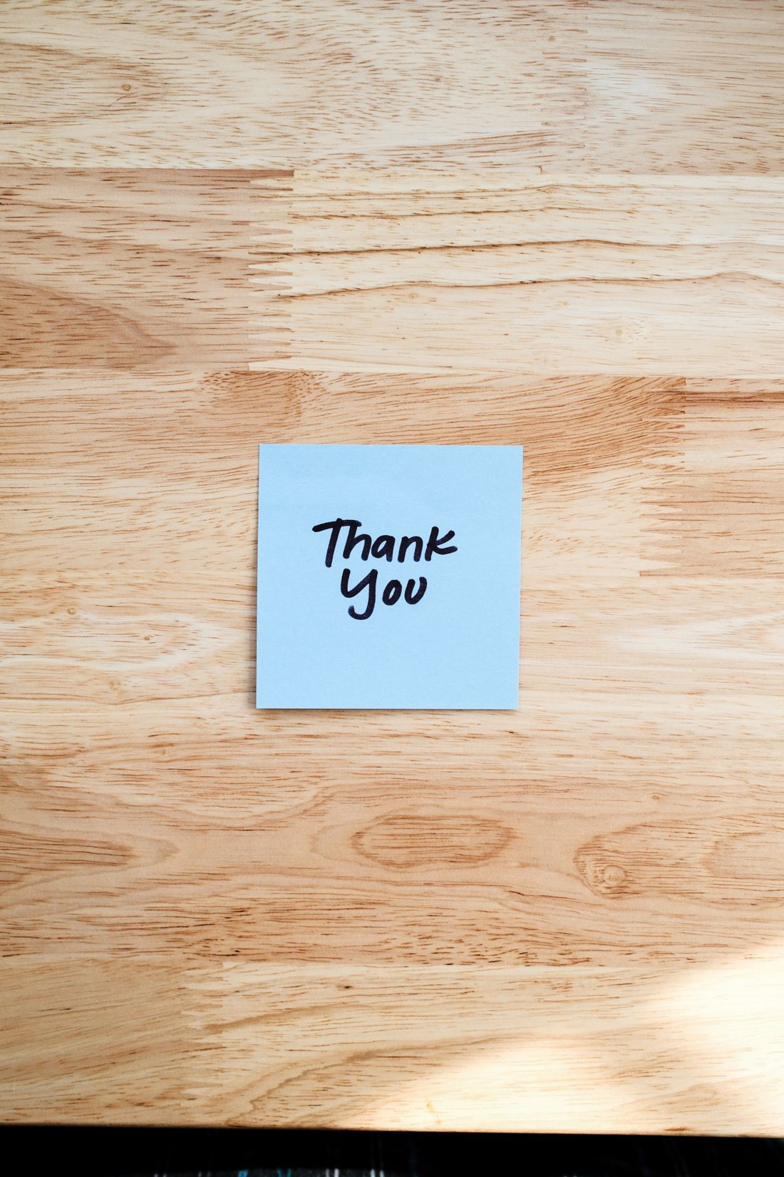 A handwritten Thank You note on a light blue sticky note resting on a wooden desk surface.