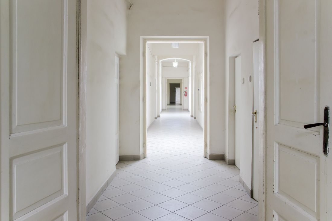 A long empty office corridor with plain white painted walls, white ceiling with recessed lighting, white doors and openings on both sides, and a hard tile floor, photographed looking down the length of the space.