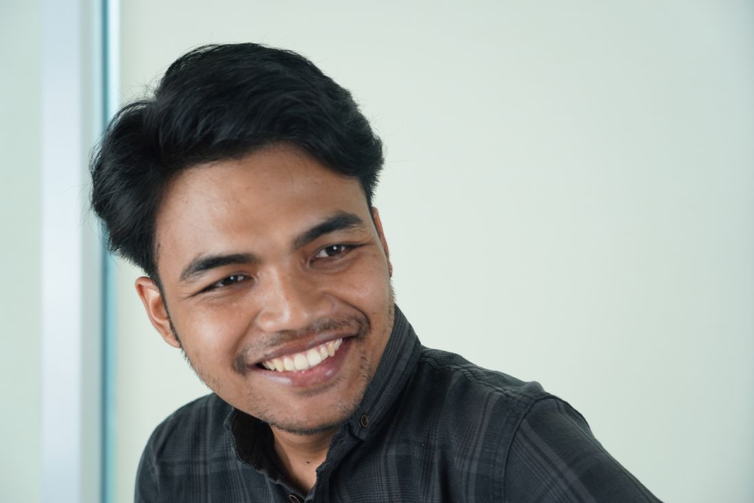 A young man in a plaid shirt smiles warmly, looking off-camera in a bright office setting.