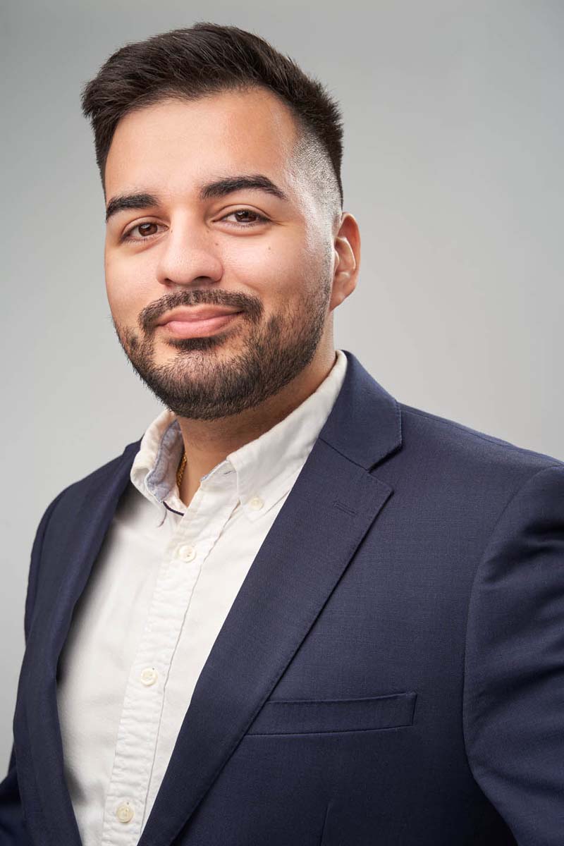 Professional headshot of a man in navy suit against light grey background.