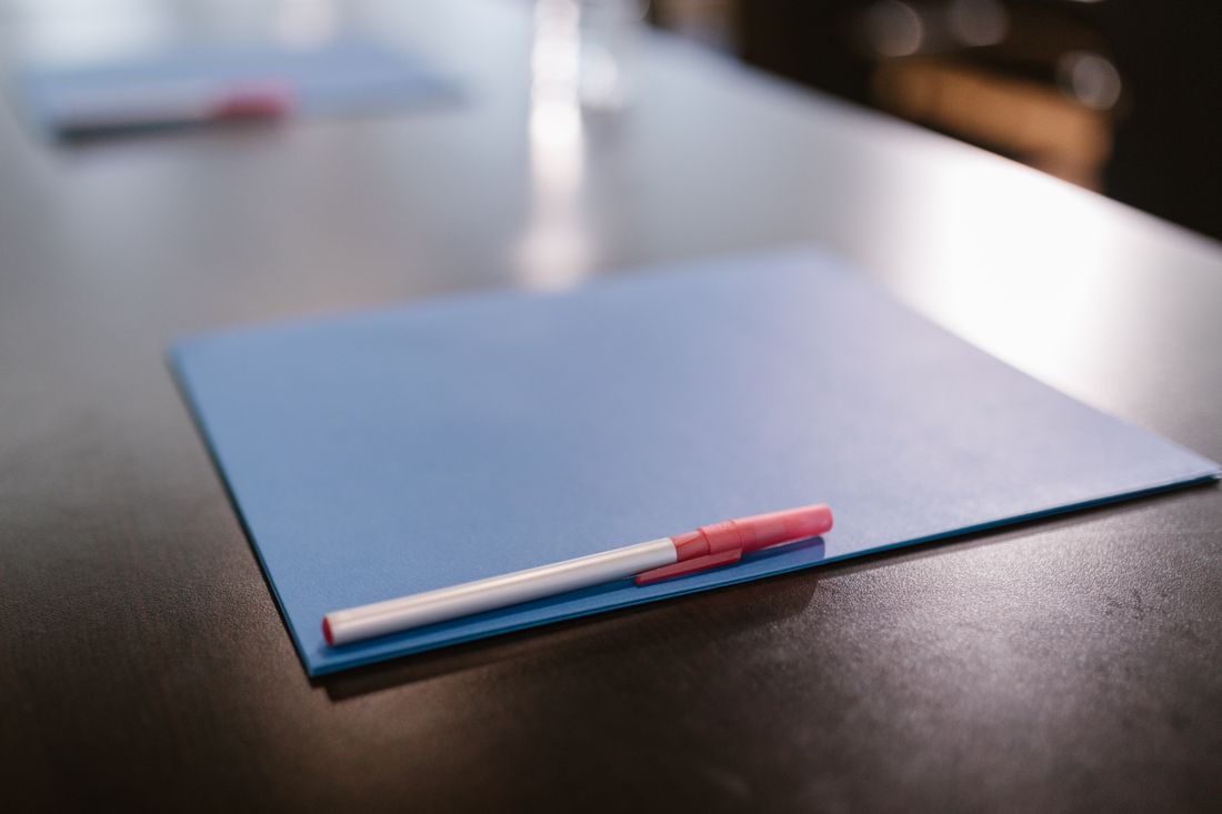 A closed blue folder with a pen resting on it, on a dark table with a second folder out of focus behind.