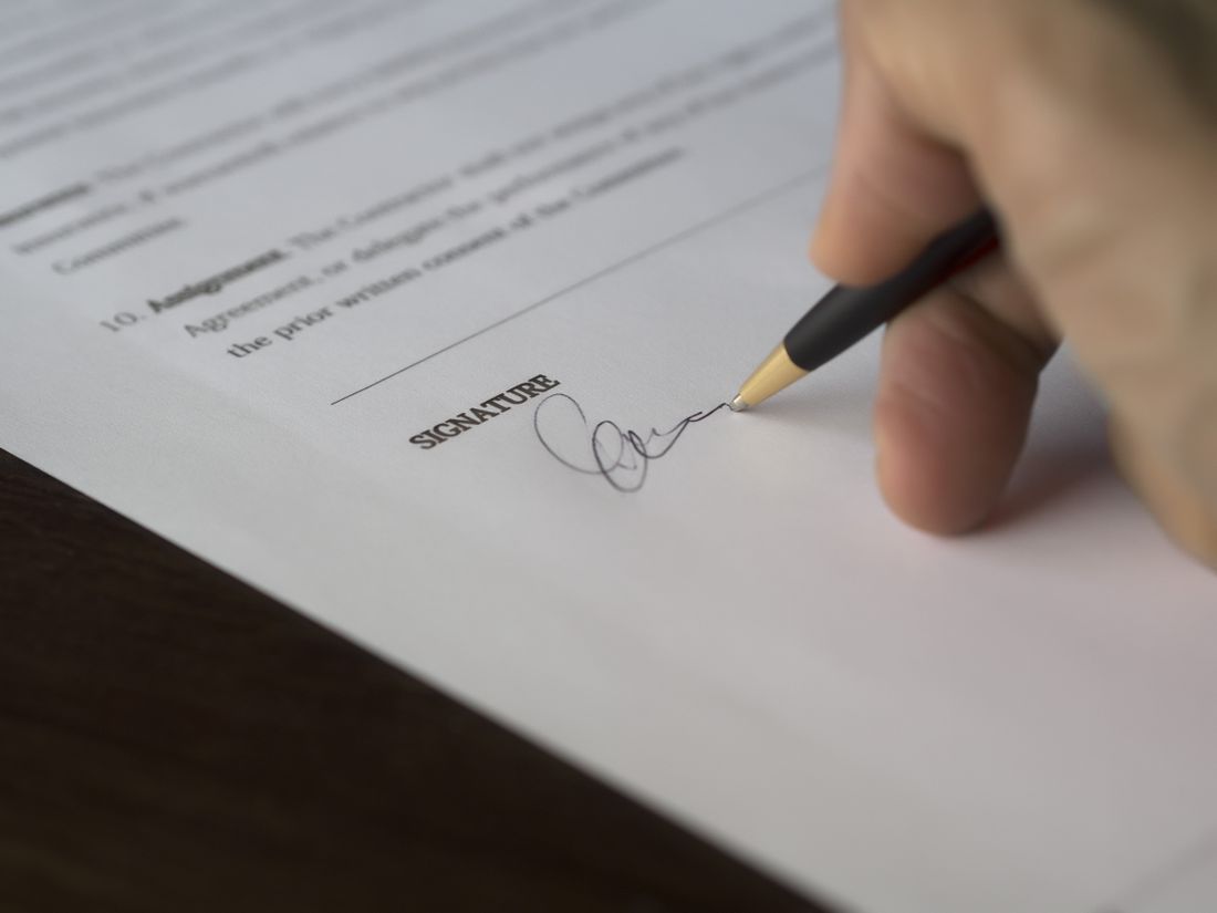 Close-up of a hand signing a contract with a pen on a wooden desk, representing a booking agreement.